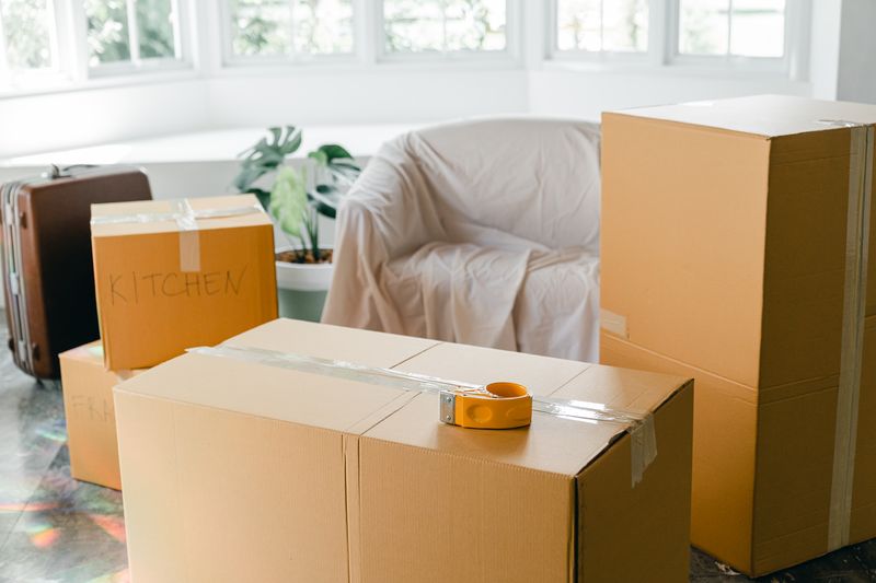 Cajas de mudanza cerradas con cinta y rotuladas en un piso luminoso, junto a un sillón tapado con una sábana