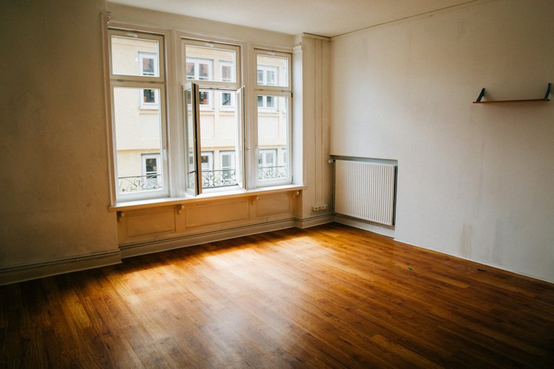 Salón vacío con suelo de madera y ventanales que dan a una calle de edificios clásicos con balcones